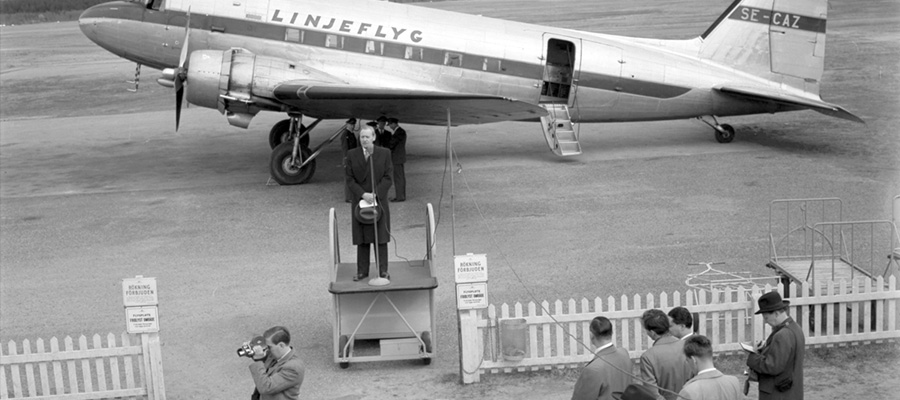 Bild. En person talar i mikrofon framför ett flygplan med texten Linjeflyg. Det finns fler åskådare i förgrunden, två med kameror.