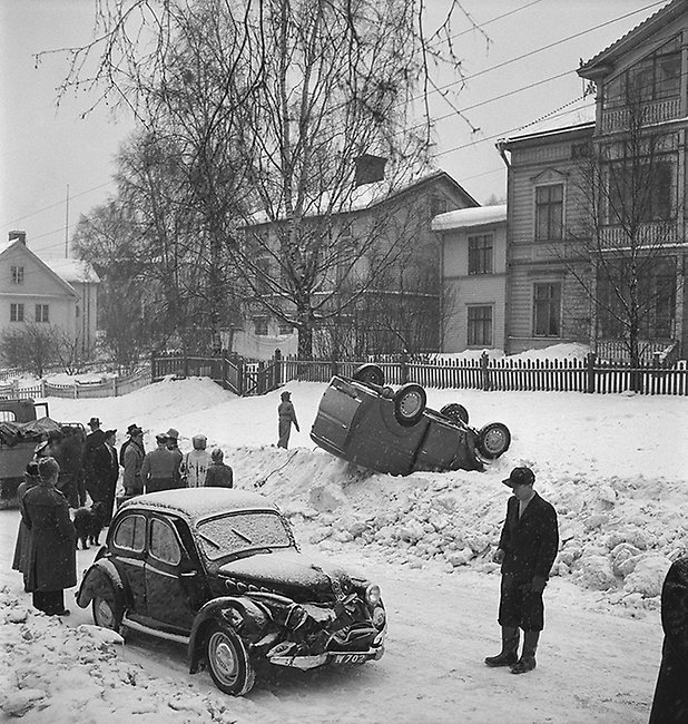 Foto. En skåpbil har slagit runt vid en trafikolycka på Bergsgatan strax väster om Esplanaden, 1951. Bilen ligger upp och ner i en snödriva längs vägkanten. Fotograf: Norrlandsbild. Bildkälla Sundsvalls museum.